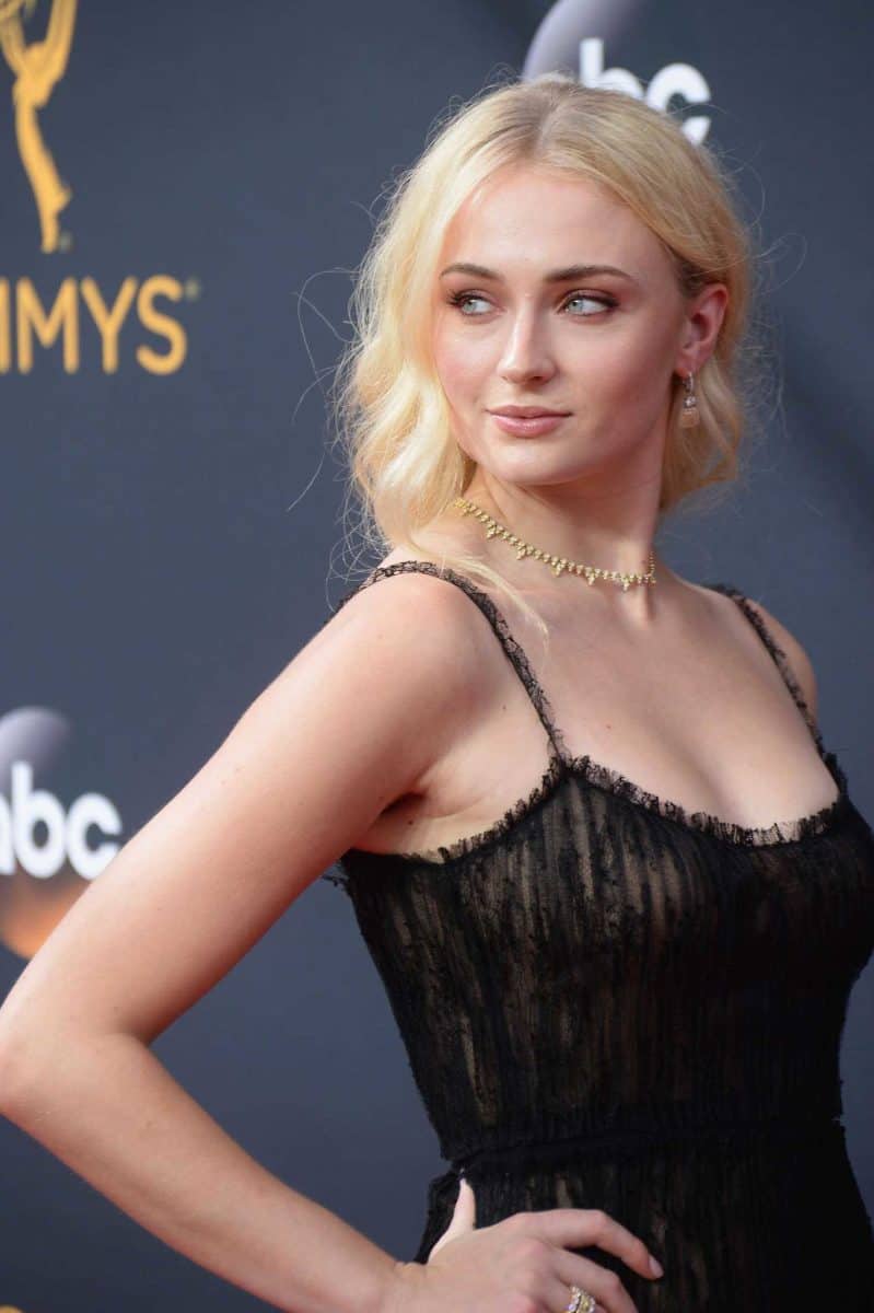 Sophie Turner with blond hair in black lace dress wears a necklace and earrings being photographed at the Emmy awards