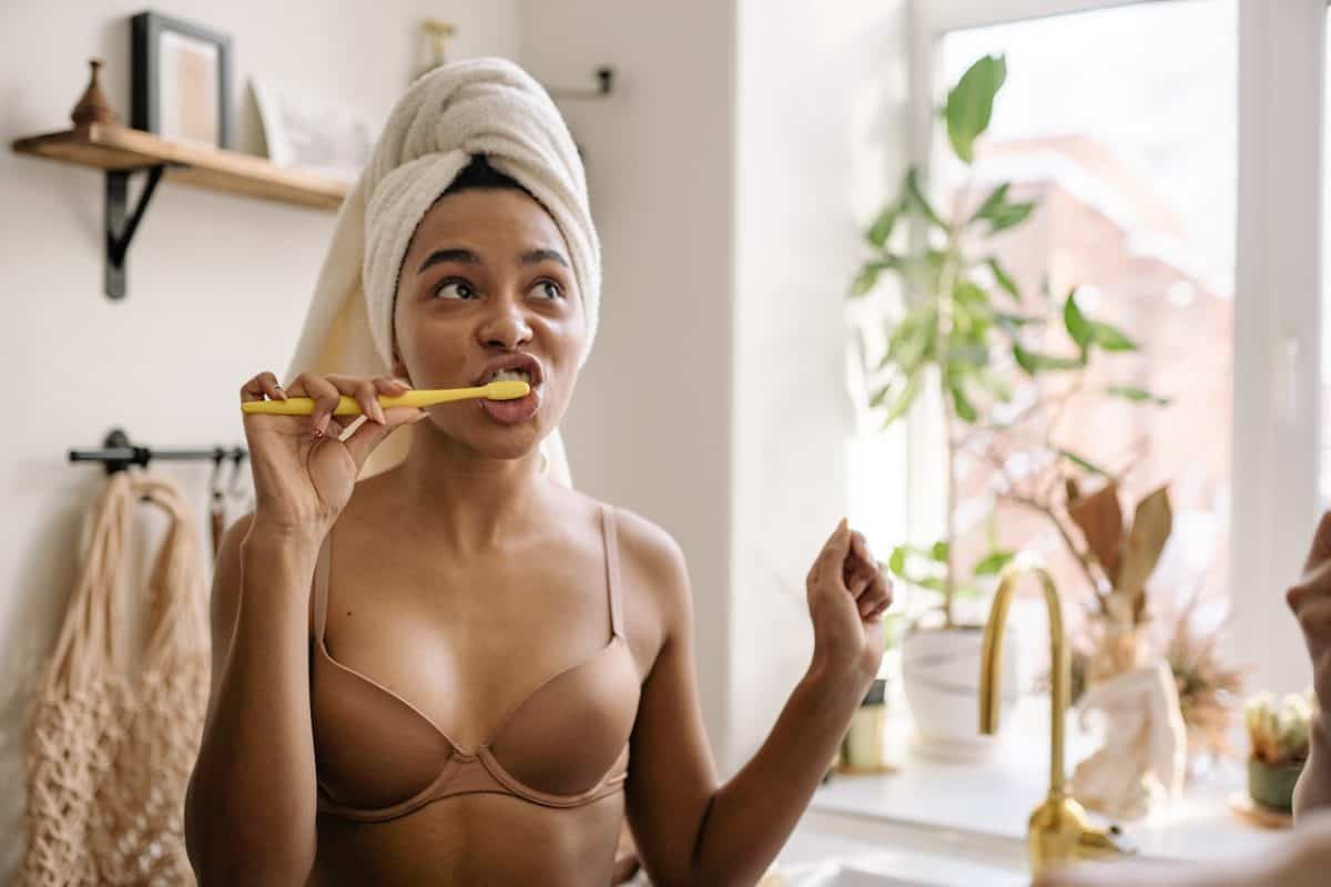 A woman wearing a brown bra brushing her teeth with a yellow toothbrush and a white towel on her head in the bathroom