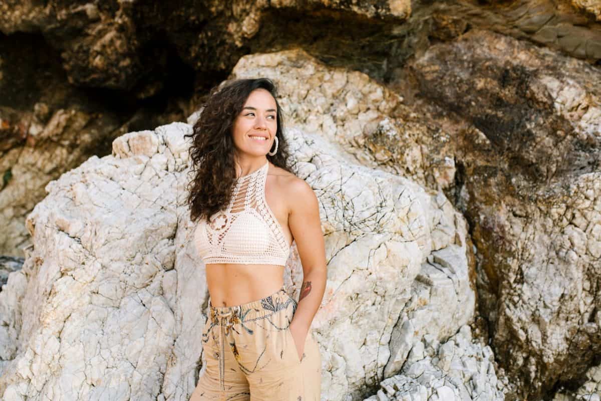A woman wearing a white knitted bralette crop top and brown shorts standing near a rocky formation