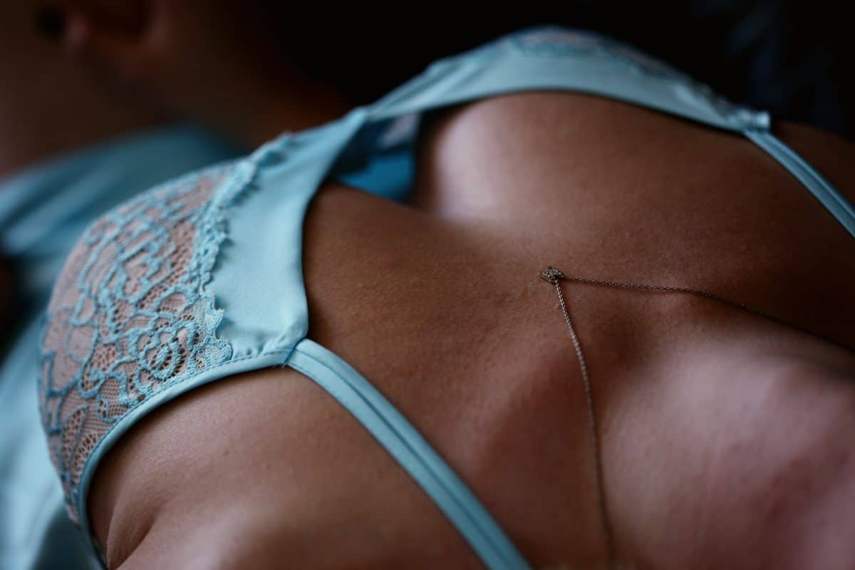 A woman wears a blue laced bra and silver necklace while lying down on a bed in a dim lit room
