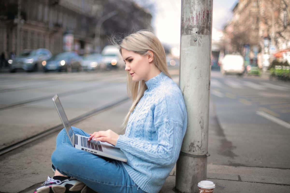 A woman wearing a light blue long sleeve shirt and blue jeans using a silver laptop while seated on the street