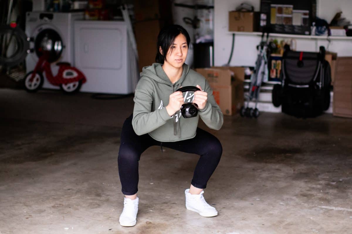 Woman wearing a green sweater and leggings doing kettlebell squats inside a garage
