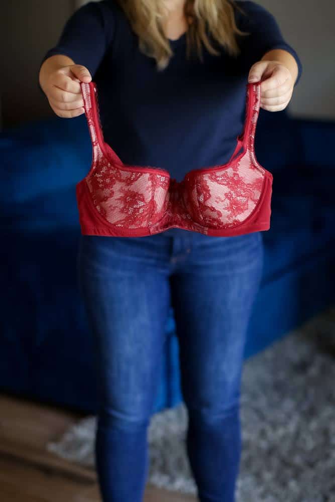 A person holds a red lace bra standing on a wooden floor near a gray carpet