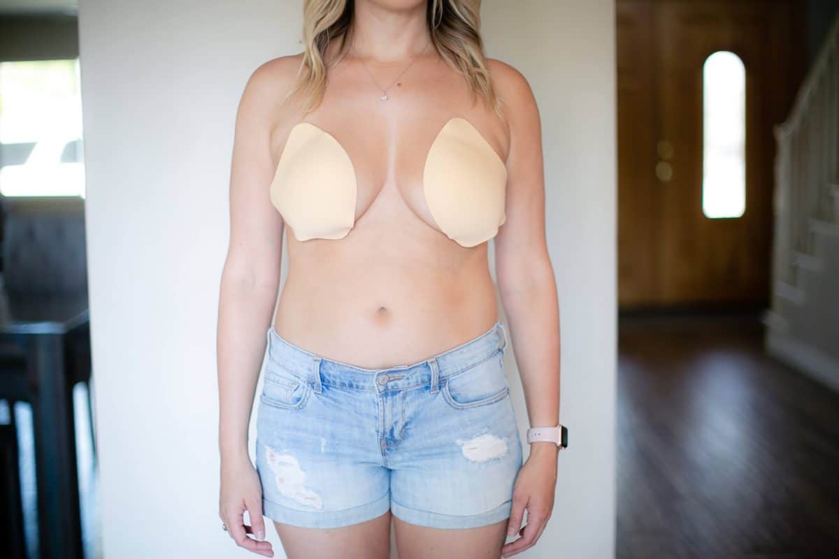 A woman wearing a nude adhesive bra and blue denim shorts standing near a white wall in the kitchen