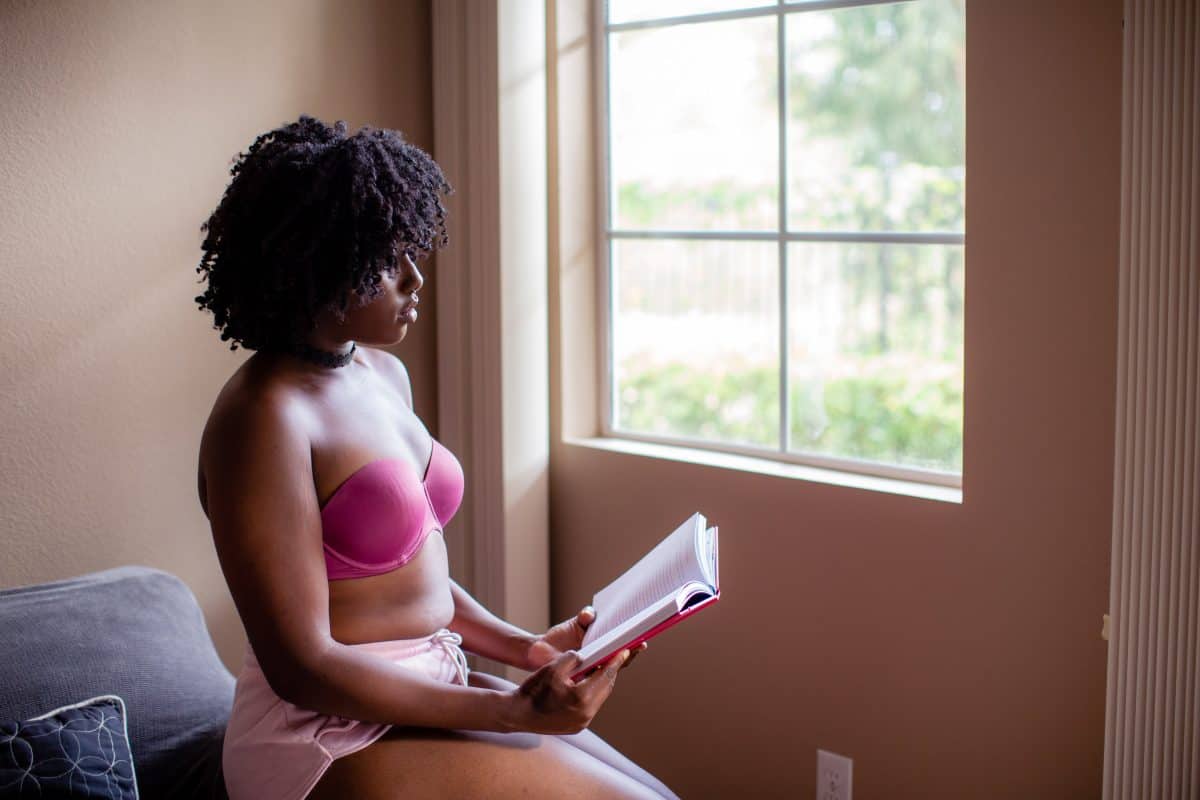 A woman wearing a pink bralette and pink shorts reads a book while seated on a gray couch