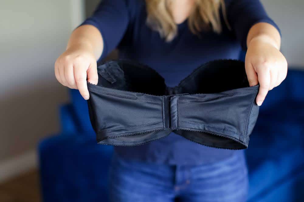 A woman wearing blue long sleeves and blue denim jeans holds a black strapless bra and is standing near a blue couch