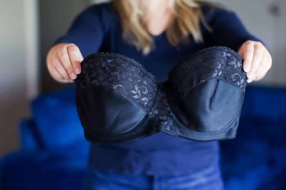A woman in long blue sleeves and blue pants holds a black strapless bra near a blue couch