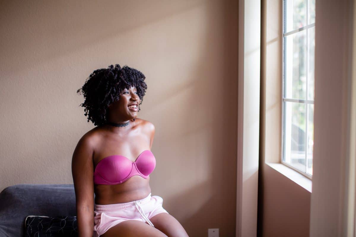 A woman wearing a pink strapless bra and pink shorts is seated on a black couch while smiling and looking out the window