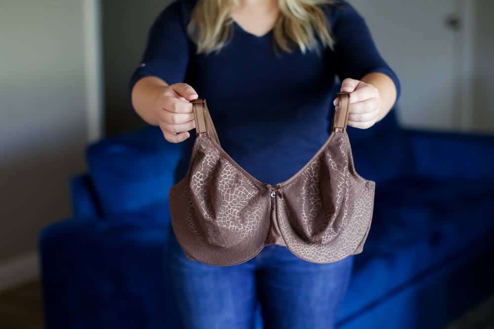 A woman in a blue blouse and blue denim jeans holds a brown minimizer bra near a blue couch