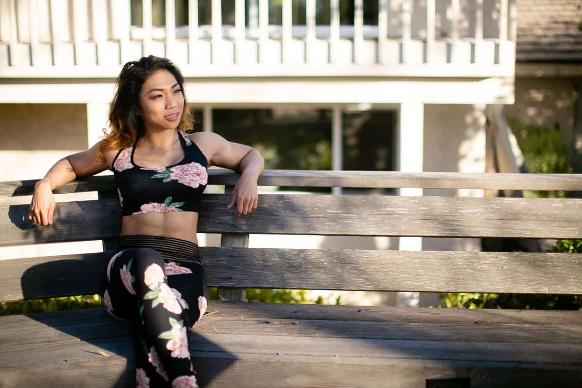 A woman wearing a black sports bra and leggings with a flower design seated on a brown wooden chair