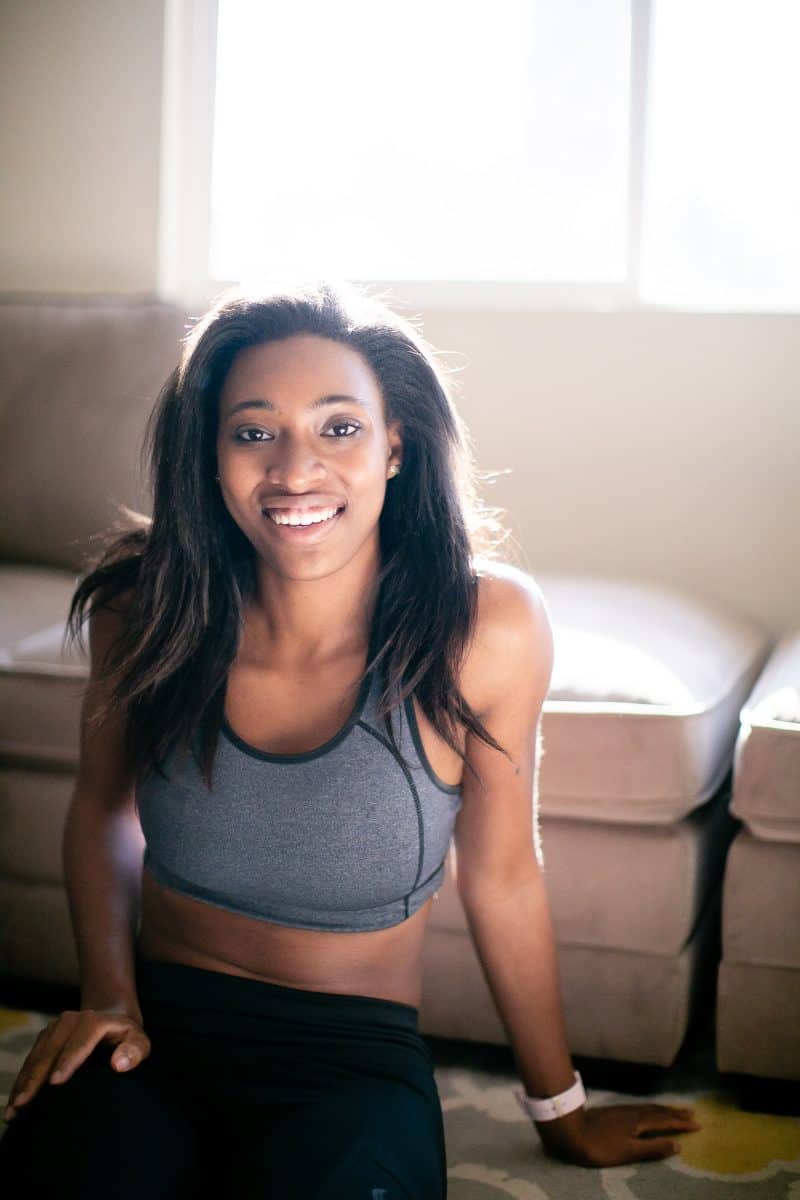 A woman in a gray sports bra and black leggings is seated on a cemented floor near a white couch