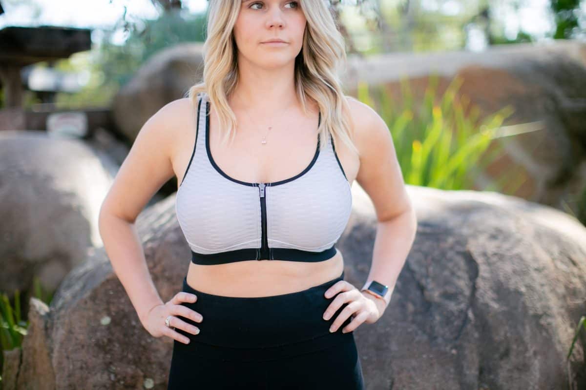 A woman wearing a gray zip-front sports bra and black leggings standing near a big rock