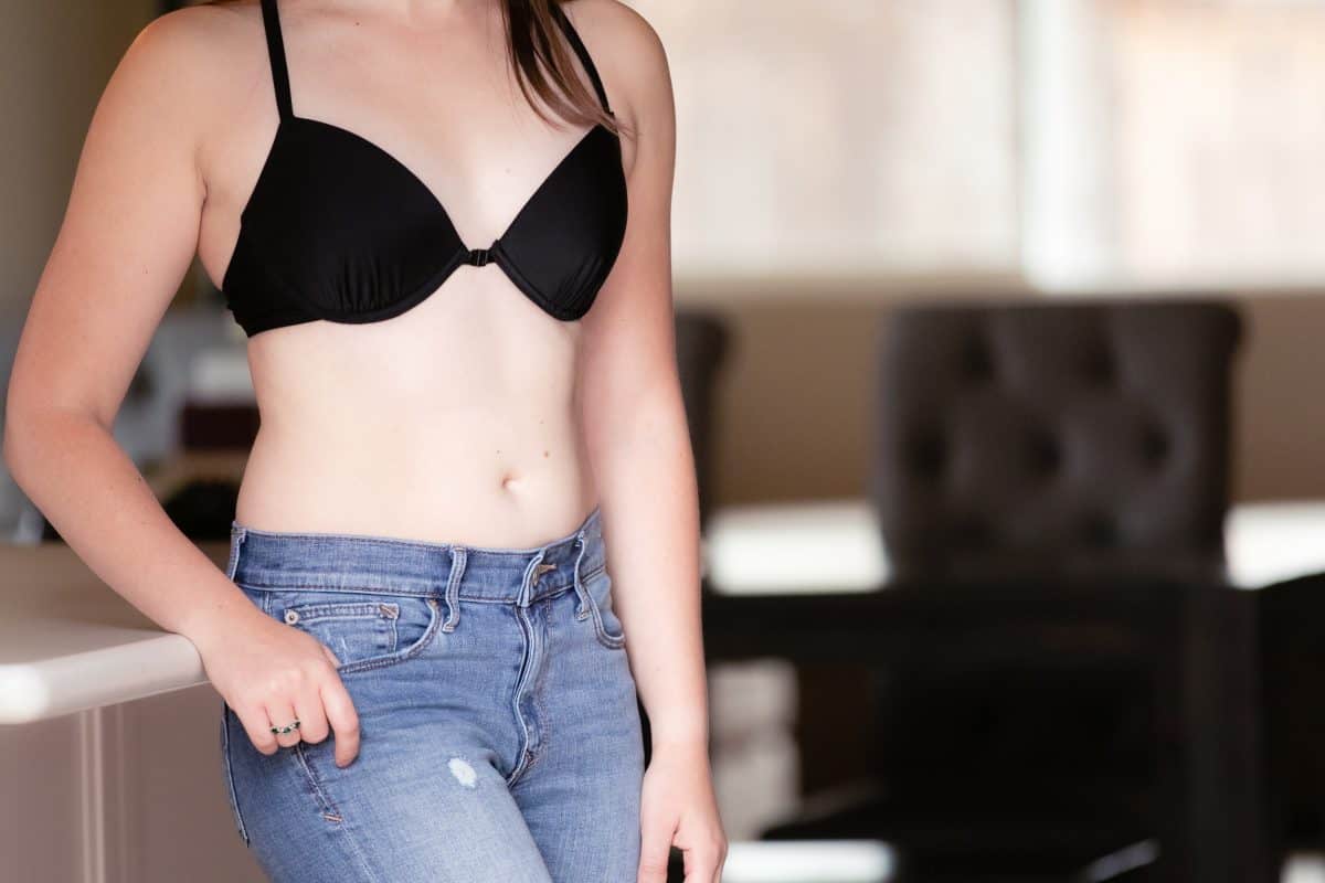 A woman wearing a black underwire bra with a front clasp and blue denim jeans leans on a white kitchen top