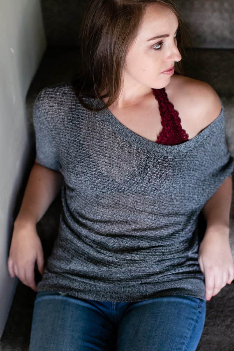 A woman wears a red bralette with a one-shoulder gray blouse and blue jeans seated on brown stairs