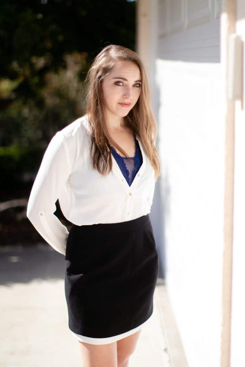 A woman in a blue bralette with a white long sleeves blouse and black mini skirt stands near a white wall on a sunny day