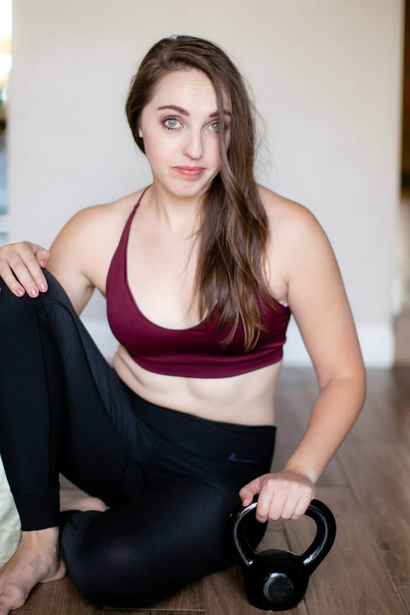 A woman in a red bralette and black leggings holds a black kettlebell on a brown wooden floor