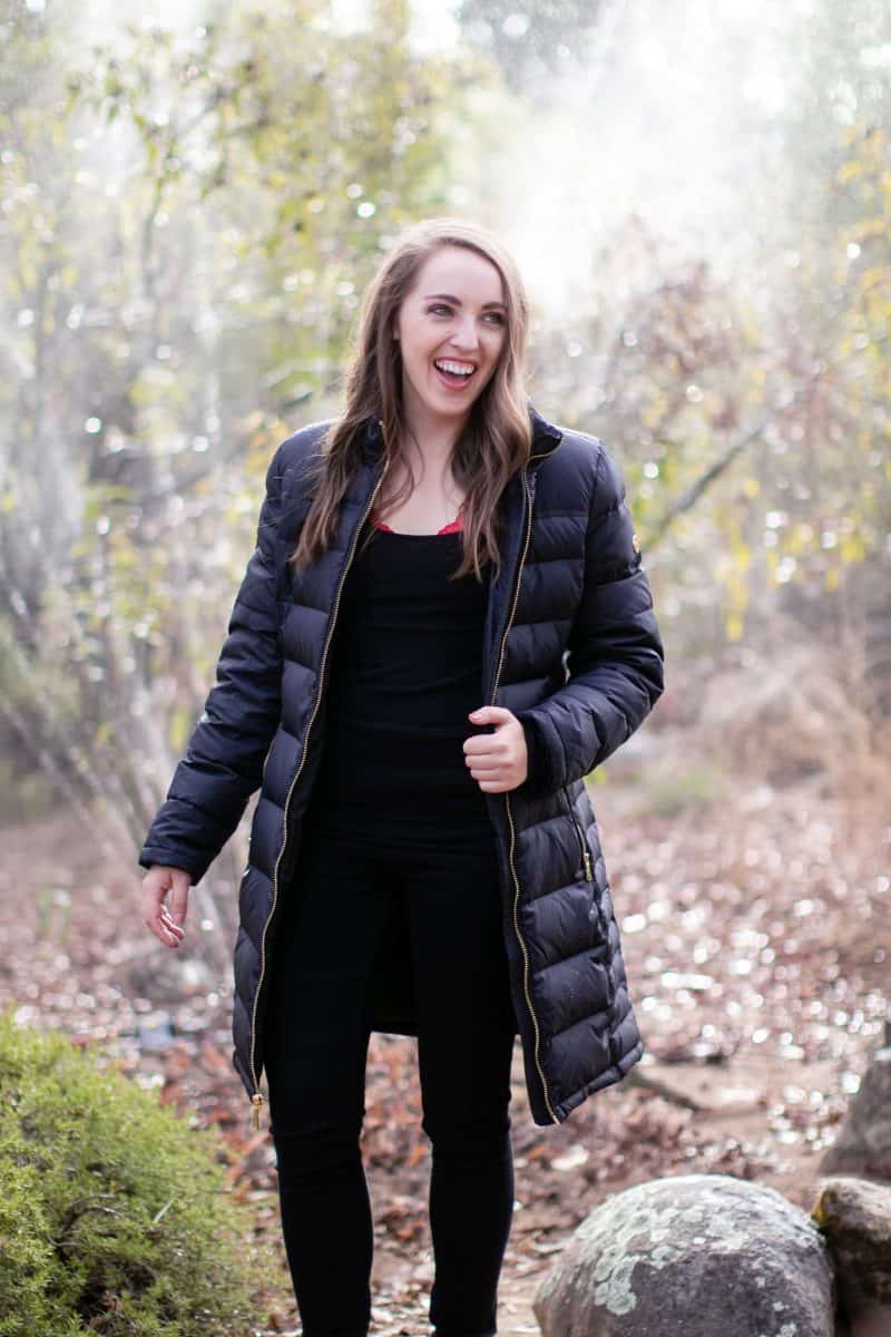 A woman in a black tank top and black padded jacket wears a black pants standing in the woods