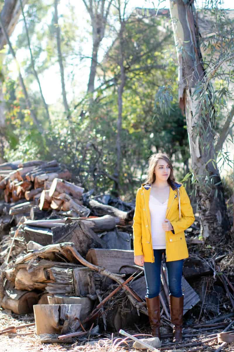 A woman in a white tank top and yellow jacket wears blue denim jeans and brown boots standing near a chopped wood.