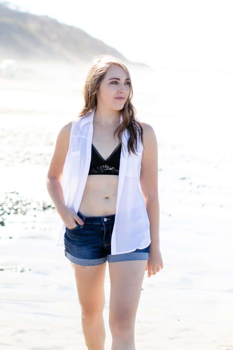 A woman wearing a black bralette with a white blouse and blue denim shorts walks on the sand on the beach