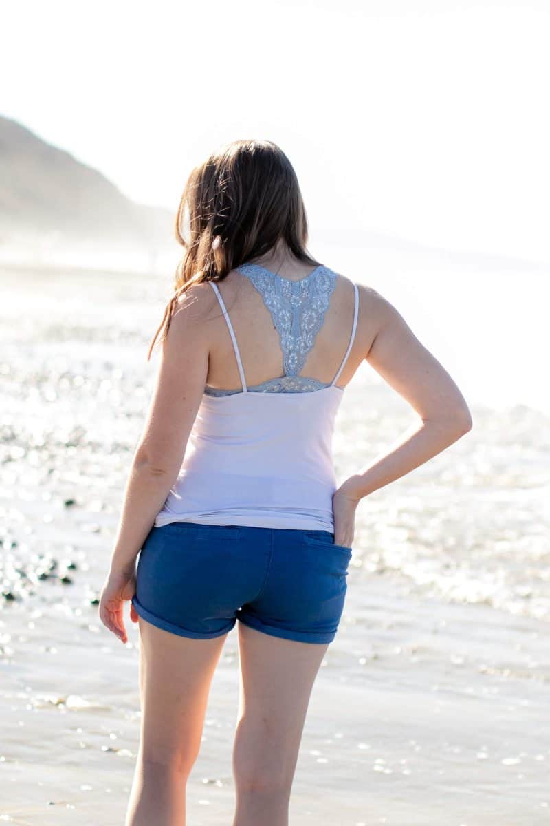 A woman facing back wearing a gray racerback bralette with a white spaghetti tank top and blue shorts standing on the sand
