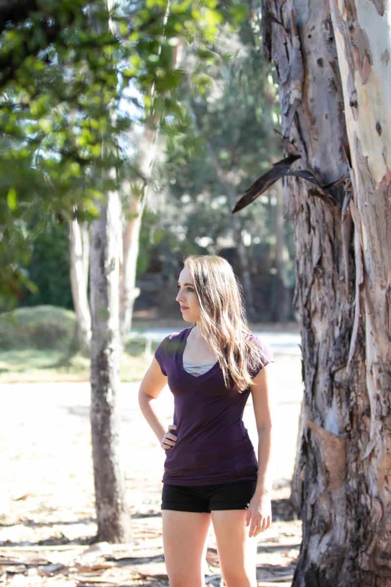 A woman wearing a gray bralette with a purple blouse and black shorts standing near a tree