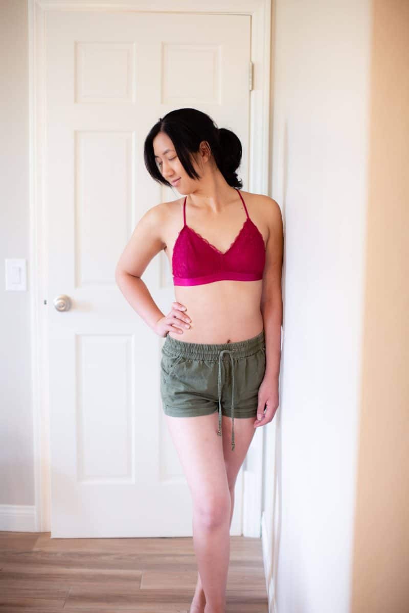 A woman in a pink lace bralette and green shorts leans on a white wall near a white door