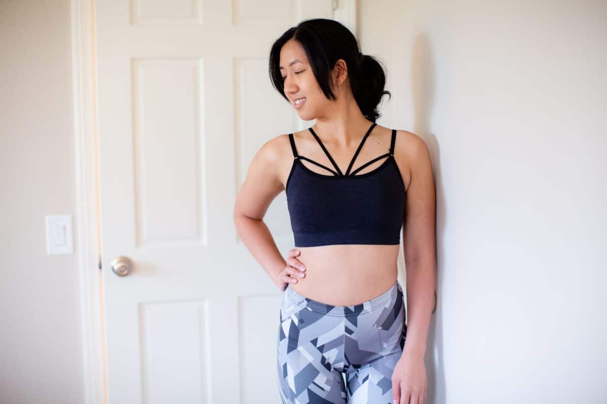A woman wearing a black bralette and printed leggings leans on a white wall in the living room