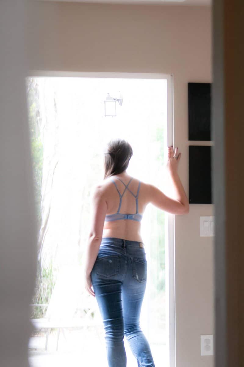 A woman who has turned her back on wears a blue bralette and blue denim jeans standing near a window in the living room