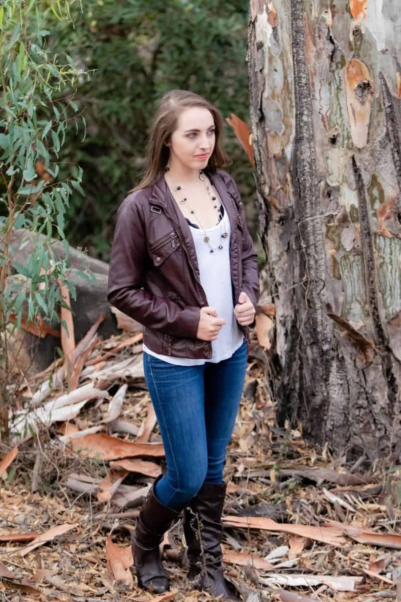 A woman in a white tank top and brown leather jacket wears blue jeans and black boots in the woods