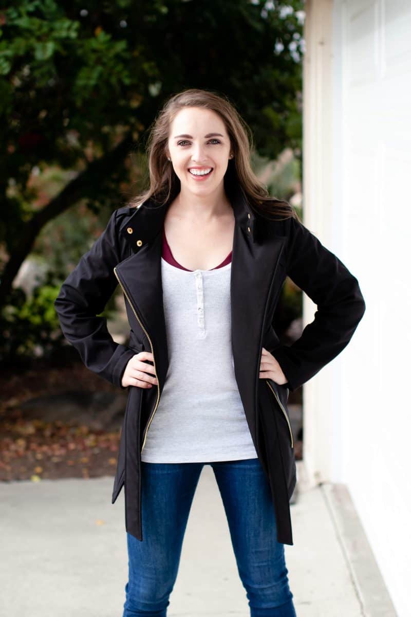 A woman in a red bralette with a white tank top wears a black jacket and blue jeans standing near a white wall