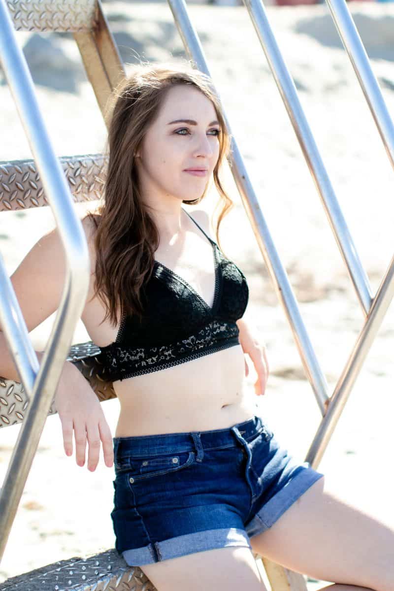 A woman wearing a black lace bralette and blue denim shorts seated on stainless stairs on the beach