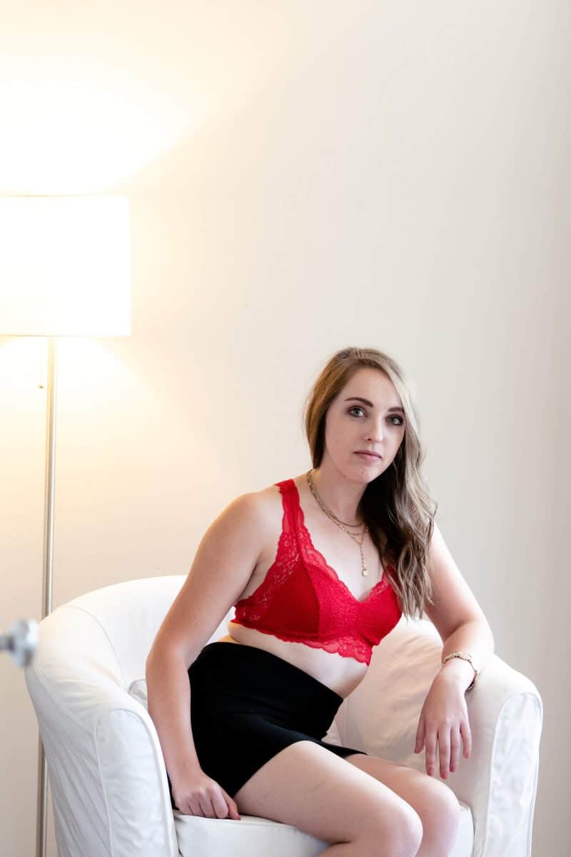 A woman wears a red lace bralette and black pencil cut skirt seated on a white couch