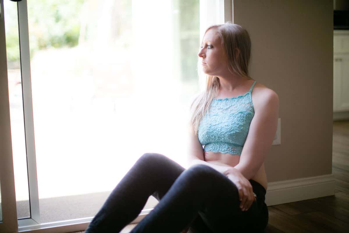 A woman wearing a blue lace bralette and black pants seated on a brown wooden floor