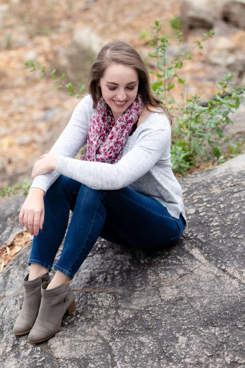 A woman in white long sleeves and blue denim jeans is seated on a big rock