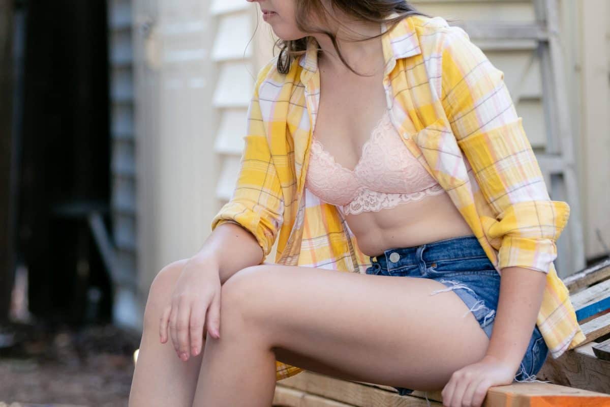 A white lace bralette with yellow checkered long sleeves and blue denim shorts worn by a woman seated on a brown wooden chair