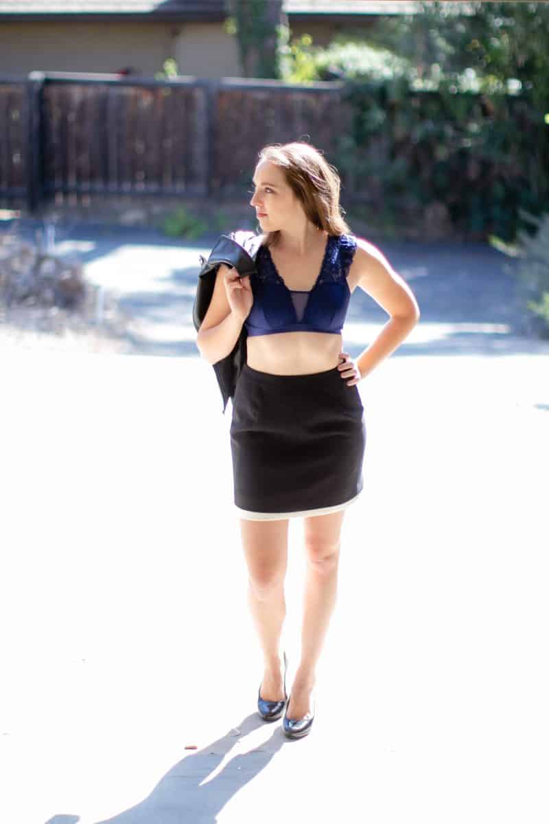 A woman wearing a blue lace bralette and black pencil cut skirt holds a black jacket standing outside on a sunny day
