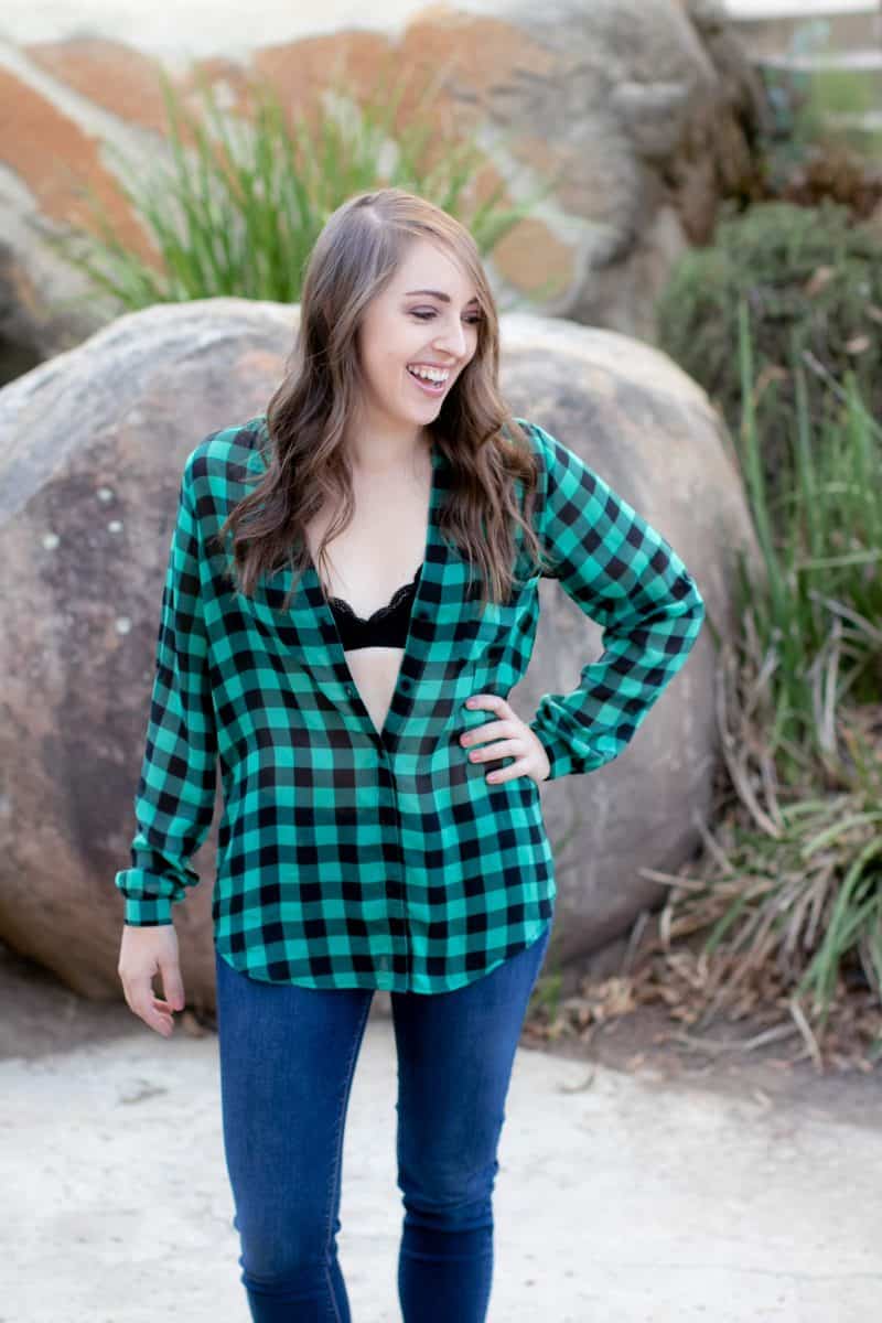 A woman wearing a black bralette with green and black checkered long sleeves and blue denim pants standing near a big rock