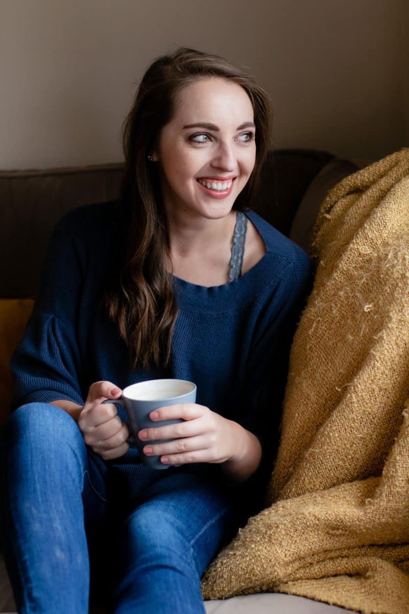 A woman in a blue blouse and blue denim jeans holds a blue mug seated on a brown couch
