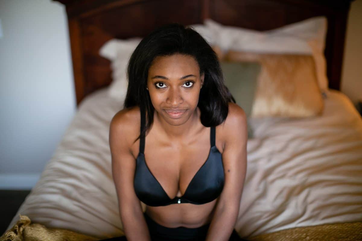 A woman wearing a black push-up bra and black leggings is seated on a white bed in the bedroom