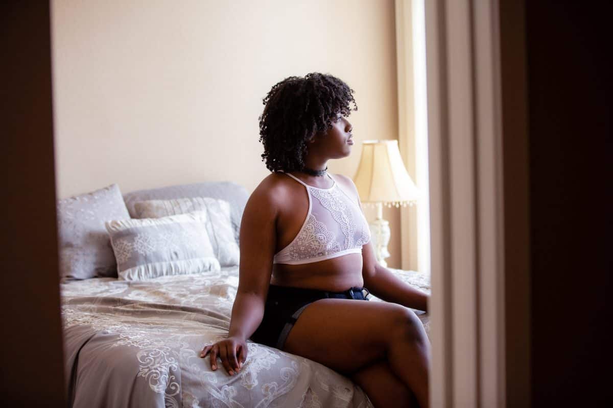 A woman wearing a white bralette and blue shorts seated on a white bed in the bedroom