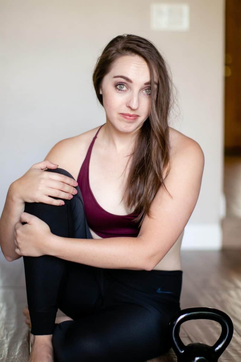 A woman wearing a red bralette and black leggings is seated on a brown wooden floor