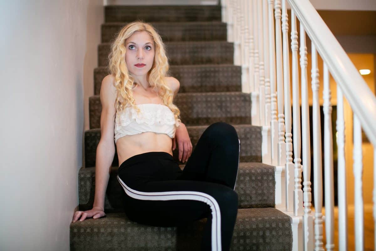 A woman is wearing a white bandeau bra and black jogging pants with white stripes on the side, seated on the stairs