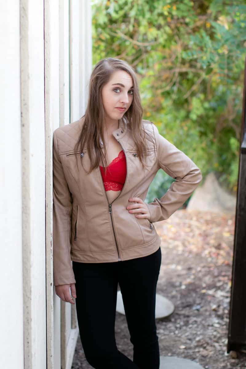A woman wears a red bralette and a brown jacket, and black pants, leaning on a white wooden wall