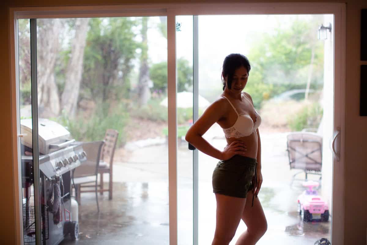 A woman wearing a white plunge bra and black shorts is standing near a sliding glass door in the living room