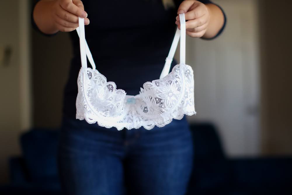 A woman wearing a black shirt and blue denim jeans is holding a white lace shelf bra and standing near a blue couch