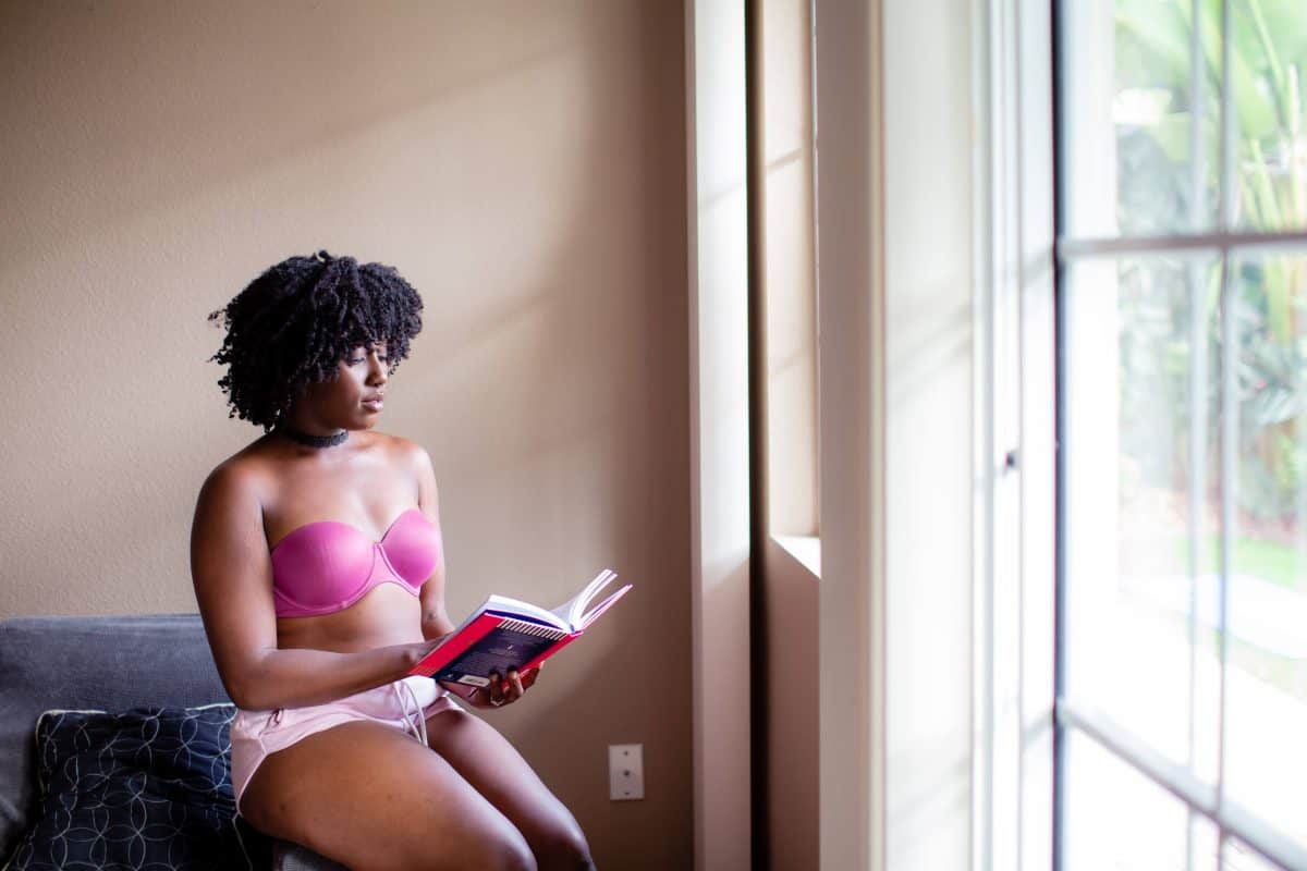 Woman in a pink strapless bra and pink shorts reading a book while seated on the arm rest of the couch