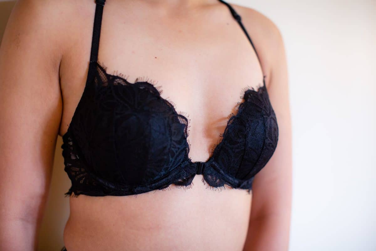 A black underwired bra with a front clasp and lace design worn by a woman leaning on a white wall