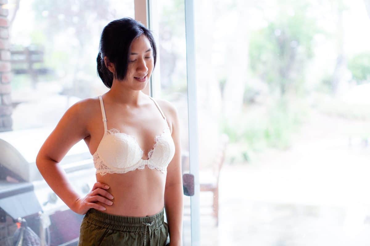 Woman in a white underwire bra with lace and front clasp wearing green shorts standing near a glass door