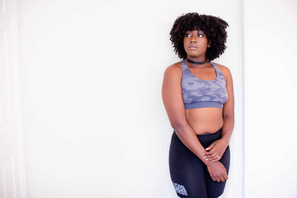 A gray sports bra with a camouflage design and black leggings are worn by a woman standing near a white wall