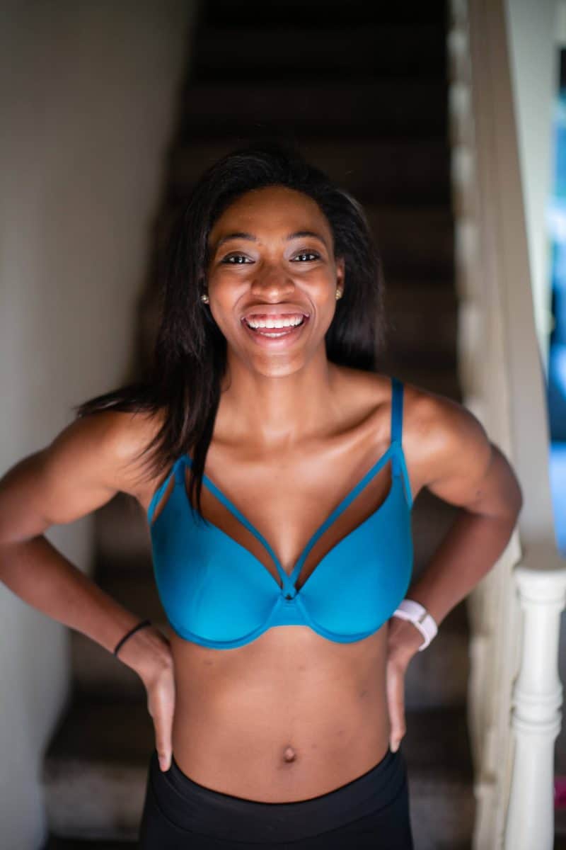 A woman who is smiling widely wears a blue push-up bra with underwire and black leggings standing near a staircase